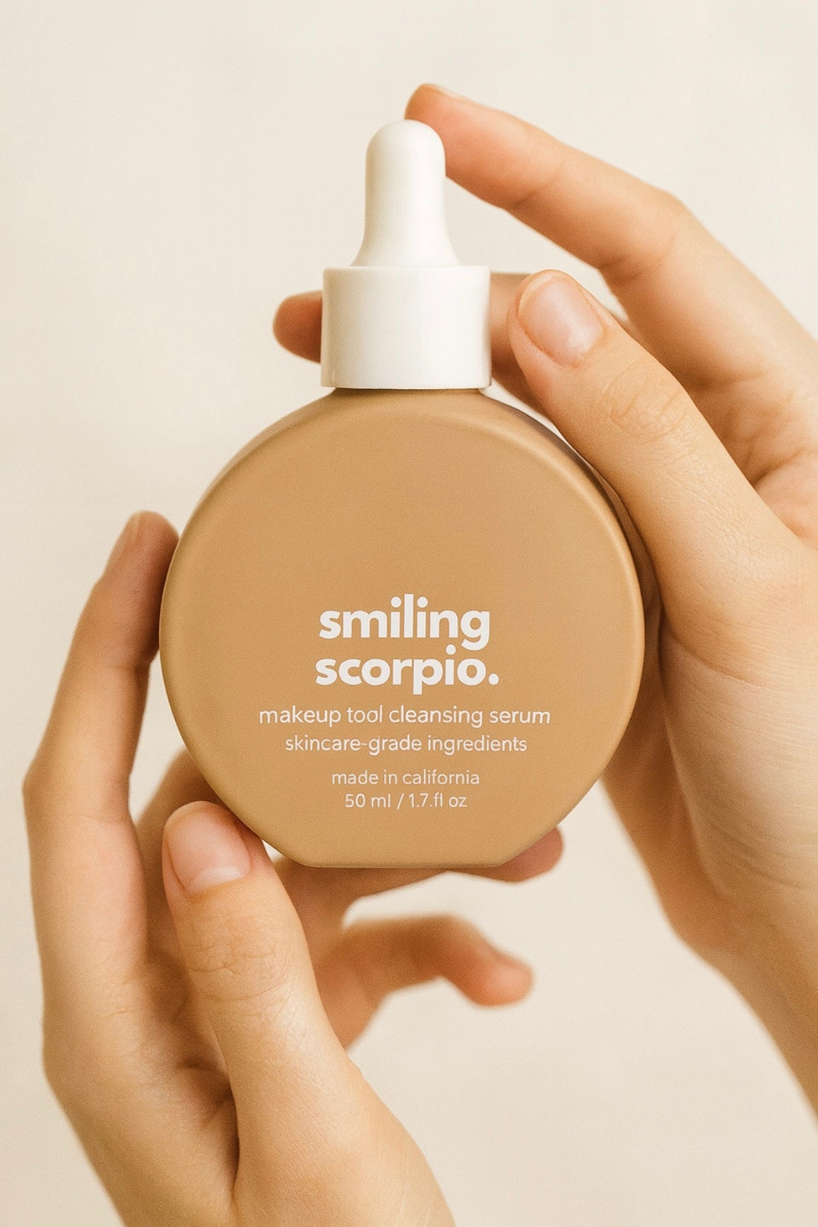 Hand holding a 'smiling scorpio' makeup tool cleansing serum bottle against a beige background